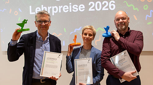 Die drei Personen stehen nebeneinander. Sie halten in der jeweils linken Hand eine Urkunde und in Kopfhöhe einen kleinen Preispokal. Dieser hat jeweils die Farbe der Fakultät: Grün, Orange und Blau.