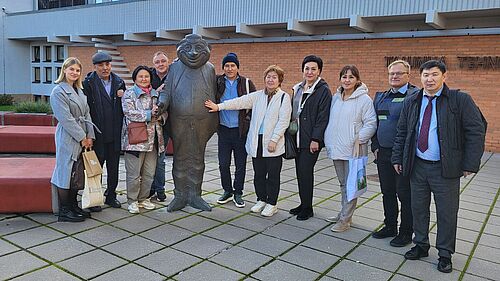 Eine Menschengruppe steht draußen. In ihrer Mitte eine Statue. Die Menschen haben Jacken an. 