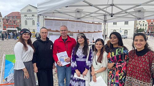 Besuch am Hochschulstand Frank Junge steht mit Dr. Anna Fomenko und fünf Studentinnen unter einem Pavillon auf dem Wismarer Marktplatz.