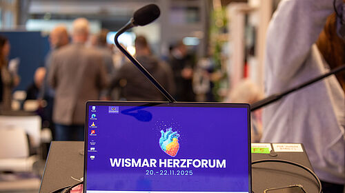 Auf dem Bildschirm eines Laptops ist eine Herzgrafik zu sehen. Dahinter im Raum sind unscharf Menschen im Gespräch zu erkennen.