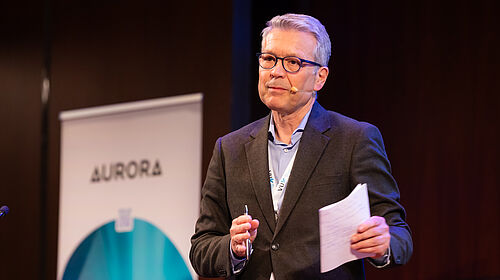 Ein Redner steht auf einer Bühne mit Headset-Mikrofon und präsentiert vor einem Aurora-Banner beim Aurora Symposium on Global Citizenship 2026.