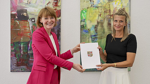 Bowen-Seidemann Die beiden Frauen stehen vor einer Wand mit zwei großen Bildern. Sie halten gemeinsam die Ernennungsurkunde in der Hand.