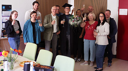 Erfolgreiche Promotion von Dominic Strube gefeiert Dominic Strube steht nach seiner erfolgreichen Promotion im Kreis von Kolleginnen und Kollegen. Die Gruppe hält Sektgläser in den Händen und feiert den Abschluss der Dissertation. Dominic Strube trägt einen Doktorhut und hält einen Blumenstrauß.