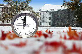 auf dem Bild zu sehen: eine Uhr vor herbstlich rot gefärbten Blättern im Schnee