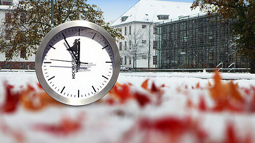 auf dem Bild zu sehen: eine Uhr vor herbstlich rot gefärbten Blättern im Schnee