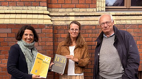 Forschung zum Umwelthaftungsrecht vorgestellt Drei Personen stehen vor einer Backsteinfassade; zwei von ihnen halten juristische Fachpublikationen in den Händen.