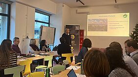 The picture shows a light room, in the middle a man is standing. He wears a dark pullover and has curls. Other participants sit and listen to him.