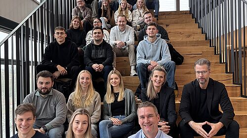 Gruppenfoto im Paulihaus Etwa 25 Personen sitzen auf einer breiten Holztreppe in einem lichtdurchfluteten Raum und lächeln in die Kamera.