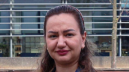 Rezvan Kakavand startet Promotion Porträt einer Frau im Freien auf dem Wismarer Campus vor einem modernen Hochschulgebäude. Sie trägt eine Jeansjacke und blickt in die Kamera.