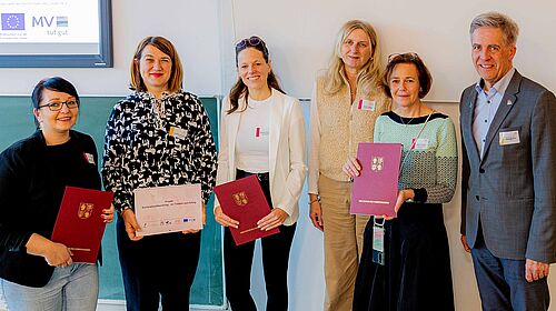 Zuwendung_2026 Fünf Frauen und ein Mann stehen nebeneinander. Sie halten Urkundenmappen mit dem Signet der Landesregierung in der Hand. Im Hintergrund ist eine Präsentation mit dem Namen der Justizministerin zu sehen.