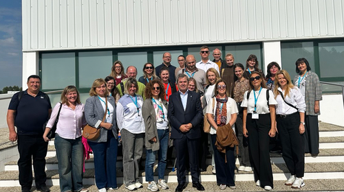 Eine Gruppe von etwa 25 Personen steht in mehreren Reihen auf einer Treppe vor einem modernen Gebäude mit Glasfassade und weißer Verkleidung. Alle tragen Namensschilder, einige mit blauen Lanyards.