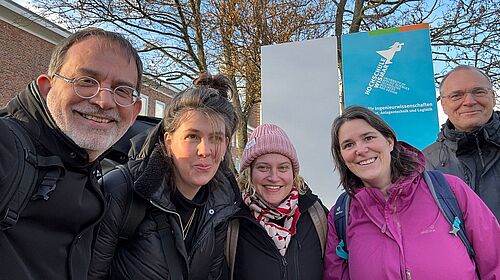 Auf dem Foto sind fünf Personen zu sehen. Links steht ein Mann mit Brille, neben ihm eine Frau mit Zopf, daneben eine blonde Frau mit rosa Mütze, daneben eine Frau mit pinker Jacke. Ganz rechts steht ein Mann mit Brille. Im Hintergrund sind Schilder vom Fachbereich Seefahrt der Hochschule Wismar zu sehen. Die Bäume im Hintergrund tragen keine Blätter.