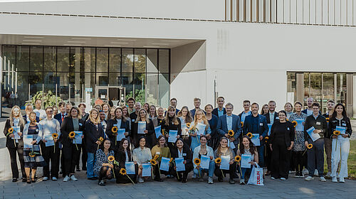 Dtl-Stip 2025 50 Personen stehen neben- und hintereinander. Einige halten Blumen und Urkunden in den Händen. Daran erkennt man die Studierenden. Im Hintergrund ist Haus 6 zu erkennen.