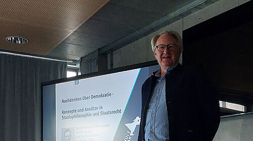 Die Person steht vor einem großen Bildschirm. Es ist der Oberkörper zu sehen und links davon eine digitale Präsentation, auf der das Logo der Hochschule Wismar zu sehen ist sowie der Titel „Nachdenken über Demokratie“.