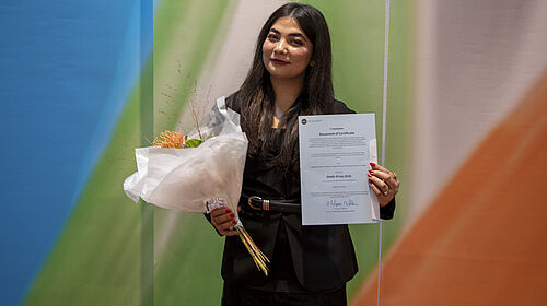 Frau in schwarzem Kleid hält Blumenstrauß und Urkunde vor einem Banner mit Schrift 'Feierliche Immatrikulation 2026'