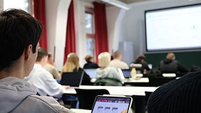 Ansicht einer Vorlesung aus der letzten Reihe. Blick auf einen Monitor und die Hinterköpfe von vor einem sitzenden Studierenden.