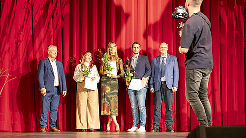 Frege-Preis 2025 Die Personen stehen auf der Theaterbühne, die rot ausgeleuchtet ist. Am rechten Rand steht ein Kameramann.